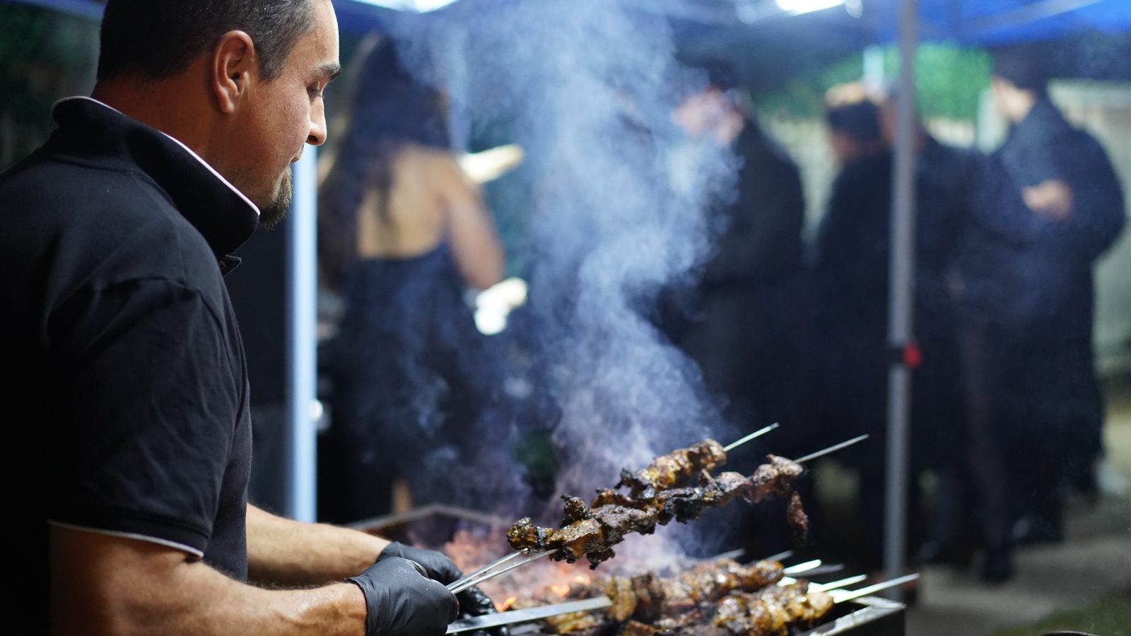 A Skewer & Spice chef turning skewers over a smoking charcoal grill while guests mingle behind