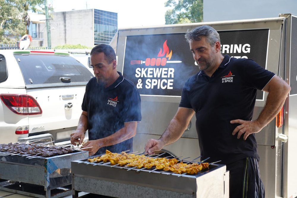 Two Skewer & Spice chefs grilling chicken skewers in front of the branded trailer